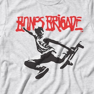 May include: A light gray t-shirt featuring the red text "BONES BRIGADE" above a black silhouette of a skateboarder in mid-air. The shirt has short sleeves and a crew neck.