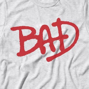 Puede incluir: Camiseta gris claro con la palabra "BAD" en letras rojas grandes, de estilo manuscrito en la parte delantera. La camiseta tiene mangas cortas y cuello redondo. La tela parece ligeramente arrugada.