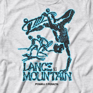 May include: A light gray t-shirt featuring a black and blue graphic of a skateboarder performing a trick. The text "LANCE MOUNTAIN" is printed below the graphic, with "POWELL PERALTA" underneath.