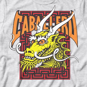 May include: A light gray t-shirt featuring a yellow dragon head with white horns and whiskers. The dragon is set against a red and black geometric background, with the word "CABALLERO" in orange above it.
