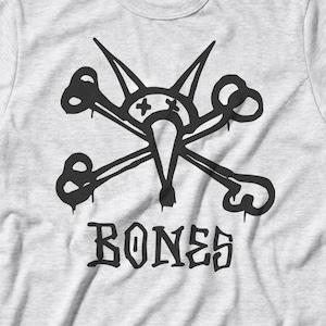 May include: A light gray t-shirt featuring a black graphic of a skull and crossbones design with a bird-like face. The word "BONES" is printed in a gothic font below the graphic. The shirt has short sleeves and a crew neck.