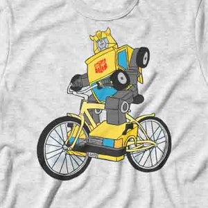 Può includere: Una maglietta grigia con un robot giallo dei cartoni animati che guida una bicicletta. Il robot indossa un casco blu e ha un camion giallo come corpo. La bicicletta ha pneumatici neri e un telaio giallo.