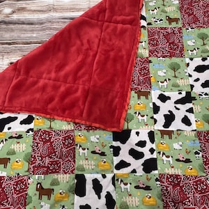 May include: A patchwork quilt featuring a red plush backing and a front side composed of squares with farm animal prints, cow spots, and red paisley patterns. The quilt displays illustrations of horses, sheep, goats, and chickens on green backgrounds.