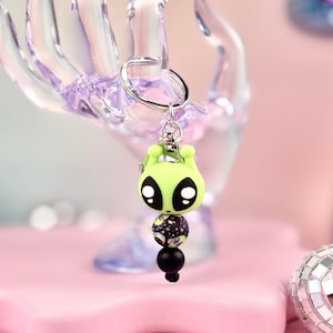 May include: A lime green alien keychain with large black eyes and a silver keyring. The alien has a rounded head and two small antennae. Below the alien is a black and patterned bead, followed by a black bead.