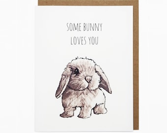 May include: A white card with brown trim featuring a hand-drawn illustration of a brown bunny with the text "Some bunny loves you".