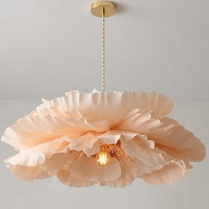 May include: A pendant light fixture shaped like a large, blooming flower with soft, pale peach-colored petals. The light hangs from a thin, braided cord attached to a small, circular gold-toned ceiling mount, featuring a single exposed light bulb.