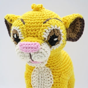 May include: A crocheted lion cub toy with yellow fur, brown ears, and a pink nose. The toy has black eyes and a white muzzle.