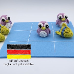 Puede incluir: Un tablero de juego de fieltro azul con un patrón de cuadrícula. Hay cinco juguetes de crochet en el tablero: tres pingüinos morados y dos peces amarillos. El tablero tiene una bandera alemana dibujada en él y el texto "pdf auf Deutsch English not yet available".