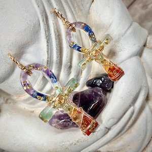 May include: Two gold-wrapped Ankh pendants with colorful, embedded stones. The pendants are on a white, textured surface with purple and green stones.