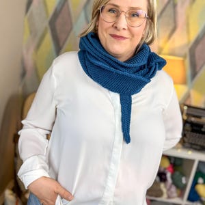 May include: A woman wearing a white button-down shirt and blue jeans is wearing a blue knitted scarf. The scarf is draped around her neck and hangs down to her waist.