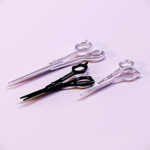May include: An assortment of professional hair styling scissors in silver and black. The scissors are arranged on a light purple surface, with two silver pairs and one black pair. The scissors are designed for precision cutting.
