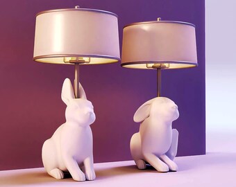 May include: Two white bunny-shaped table lamps with beige lampshades. The lamps have a gold-colored trim on the lampshades and a gold-colored stem. The lamps are sitting on a light purple surface with a dark purple background.
