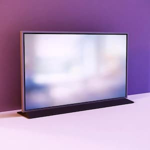 May include: A modern flat-screen television with a silver frame and a black base. The screen displays a blurred, colourful image. The TV is set against a purple wall and a white surface.