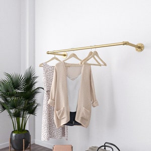 Modern minimalist interior with a golden wall-mounted clothes rack holding a beige sweater and two wooden hangers.