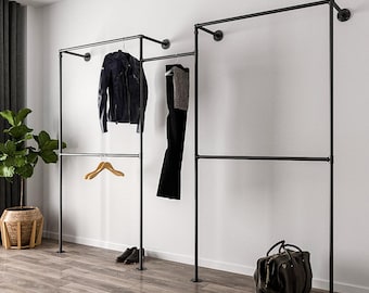 Op de afbeelding: Zwarte metalen kledingrekken met hangende kleding en accessoires. Een zwarte leren jas, een zwarte broek en een grijze trui hangen aan de rekken. Een houten kleerhanger, zwarte schoenen en een donkergroene weekendtas zijn ook zichtbaar.