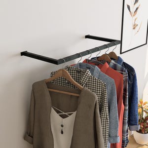 Puede incluir: Un perchero de metal negro montado en la pared exhibe una variedad de prendas. Una blazer color beige con una parte superior blanca y un colgante cuelga de una percha de madera. Otras camisas incluyen cuadros, roja y una chaqueta vaquera. Se ve una impresión botánica enmarcada.