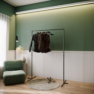 Op de afbeelding: Een moderne interieurscène met een zwart kledingrek met verschillende kledingstukken aan houten hangers. Een groene fluwelen stoel en een rond tapijt staan op de voorgrond. De muren zijn groen en wit geschilderd.
