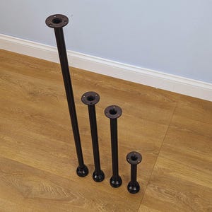 Puede incluir: Cuatro patas de mesa de metal negro de diferentes alturas, cada una con una placa de montaje redonda en la parte superior y un pie decorativo. Las patas están dispuestas en diagonal sobre una superficie de vetas de madera, con una pared azul claro al fondo.