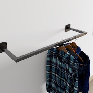 Może przedstawiać: Ciemnoszary metalowy wieszak na ubrania zamontowany na białej ścianie. Wieszak mieści trzy drewniane wieszaki z niebieską koszulą w kratę i dżinsową kurtką. Wieszak ma prosty, nowoczesny design.