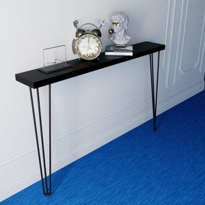 Puede incluir: Una consola negra con patas de horquilla, que sostiene un despertador vintage, un busto y un libro pequeño. La mesa está colocada contra una pared blanca y una alfombra azul. El reloj tiene un marco plateado y una esfera blanca.