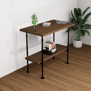 Puede incluir: Un escritorio de madera marrón con un estante inferior, sostenido por tuberías de metal negro. Una pequeña jarra con vegetación se encuentra encima, junto con libros y una revista. Una planta en maceta está en el fondo.