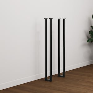 Puede incluir: Tres patas de mesa de metal negro con perfil cuadrado. Las patas están colocadas contra una pared blanca y un suelo de madera marrón. Las patas están diseñadas para la construcción de muebles.