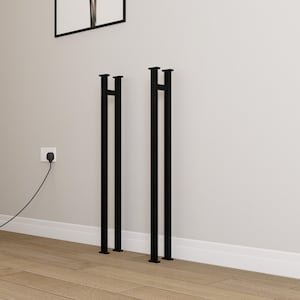 Puede incluir: Dos patas de mesa de metal negro con un diseño minimalista. Cada pata tiene una base rectangular y una parte superior con una barra horizontal. Las patas están colocadas contra una pared gris claro y un suelo de madera.