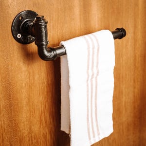 Op de afbeelding: Een zwarte industriële pijpen handdoekhouder gemonteerd op een houten oppervlak. Een witte handdoek met drie beige strepen hangt aan de houder. De houder is gemaakt van zwarte metalen buizen en fittingen.