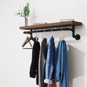 Op de afbeelding: Een aan de muur gemonteerde kledingrek met een donkerhouten plank en zwarte metalen pijpaccenten. Het rek bevat diverse kledingstukken, waaronder een zwarte jas, een geruit overhemd en een spijkerjas. Een kleine plant en boeken staan op de plank.