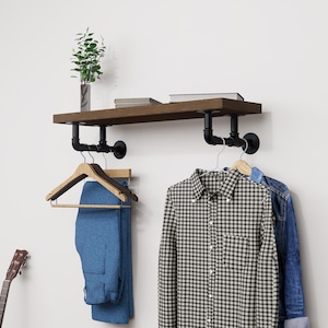 Könnte beinhalten: Ein rustikales Wandregal mit dunkler Holzoberfläche, getragen von schwarzen Rohrfittings. Darunter hängen ein kariertes Hemd, eine Jeansjacke und eine blaue Hose an Holzbügeln. Eine Gitarre lehnt an der Wand.