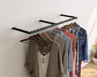 May include: A black metal clothing rack mounted on a white wall, displaying various garments on wooden hangers. The clothes include a brown cardigan, a white top, checkered shirts, and a denim jacket.