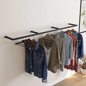May include: A black metal clothing rack mounted on a white wall, displaying various garments on wooden hangers. Items include a denim jacket, a tan blazer, a plaid shirt, and a gray sweater. A framed botanical print and potted plants are also visible.