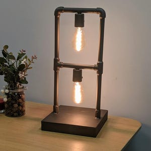 Op de afbeelding: Industriële tafellamp met een donker metalen frame en twee Edison-stijl gloeilampen. De lamp, geconstrueerd met pijpfittingen, staat op een vierkante basis. Een kleine glazen pot met decoratieve elementen en een kaars staan op de tafel.