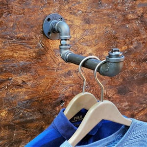 Op de afbeelding: Industriële wandkledingrek gemaakt van gegalvaniseerde stalen buizen en fittingen. Het rek heeft een flens voor montage, een gebogen pijpsectie en een haak om kleding op te hangen. Twee houten kleerhangers houden blauwe shirts vast.
