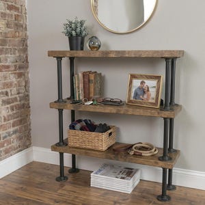 Può includere: Una scaffalatura a tre livelli con ripiani in legno marrone scuro sostenuti da tubi metallici industriali neri. Gli scaffali ospitano una pianta in vaso, una cornice, un cesto intrecciato e una pila di riviste per riporre vari oggetti.