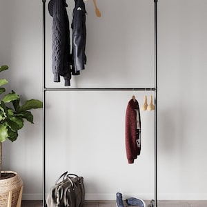 May include: A black metal clothes rail with two horizontal bars and vertical supports. A dark blue cable-knit sweater and a black leather jacket hang from the top bar. A burgundy jacket hangs below. A duffel bag and trainers sit below.