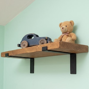 May include: A rectangular wooden shelf with a natural grain finish, supported by two black metal brackets. A dark blue wooden toy car with large wheels and a light brown plush teddy bear with a small ribbon are placed on the shelf surface.