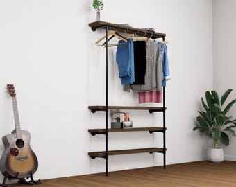 Op de afbeelding: Een zwart metalen en houten kledingrek met hangende kleding en planken. Het rek heeft een bovenste plank met een kleine plant en een gitaar leunt tegen de muur. Kleding omvat een blauwe spijkerbroek, een zwart shirt en een geruit shirt.