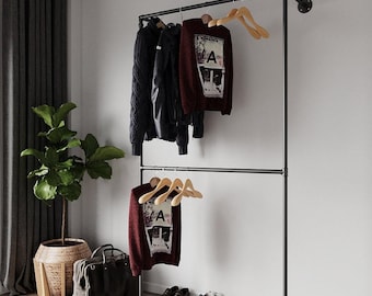 Op de afbeelding: Een zwart metalen kledingrek met twee horizontale stangen, met diverse kledingstukken op houten hangers. Een bordeauxrode trui met de letter "A" is zichtbaar. Andere items zijn een zwarte leren jas, een zwarte tas, schoenen en een potplant.