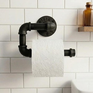 Puede incluir: Portarrollos de papel higiénico de tubería industrial negra montado en una pared de azulejos blancos. El soporte presenta una placa de montaje redonda con el número "1914" y un rollo de papel higiénico blanco.