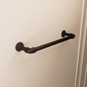 Puede incluir: Una barra de toalla negra de estilo industrial montada en una pared color crema. La barra está hecha de tuberías y accesorios de metal oscuro, con placas de montaje circulares en cada extremo. El diseño sugiere una estética rústica o moderna.