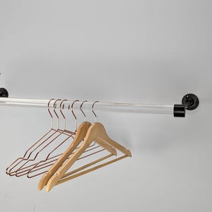 May include: A clear acrylic clothing rod mounted on a white wall with black metal hardware. Several hangers are displayed, including copper-colored metal and natural wood hangers. The rod is designed for hanging clothes.