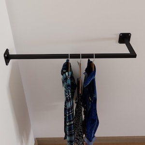 May include: A black metal clothes rail fixed to a white wall. Several garments hang from the rail, including a blue and green plaid shirt and a dark blue denim jacket. The rail has a simple, modern design.