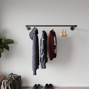 May include: A black metal clothes rail fixed to a light grey wall. A dark blue cable knit sweater, a black leather jacket, and a burgundy sweater hang from the rail. Four pairs of black shoes are on the floor.