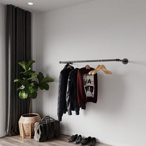May include: A modern interior scene featuring a clothing rack with a black cable-knit sweater, a black leather jacket, and a burgundy sweater with a graphic. A large gray bag, shoes, and a potted plant complete the room's decor.