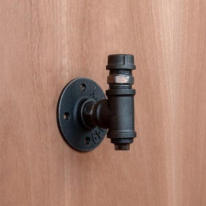 Op de afbeelding: Een zwarte industriële pijpfitting wandhaak. De haak is gemaakt van metaal en heeft een ronde basis met schroefgaten. De pijpfitting is bevestigd aan de basis en heeft een T-vorm met een schroefdraadeinde. De achtergrond is een lichtbruin houten oppervlak.