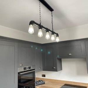 Puede incluir: Lámpara colgante industrial negra con cinco bombillas expuestas. La lámpara está suspendida por cadenas y tiene un diseño de tubería. Los armarios de la cocina son grises.