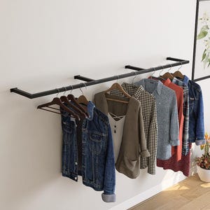 Könnte beinhalten: Eine schwarze Metall-Kleiderstange an einer weißen Wand, die verschiedene Kleidungsstücke auf Holzbügeln zeigt. Zu den Artikeln gehören eine Jeansjacke, ein beiger Blazer, ein kariertes Hemd und ein grauer Pullover. Ein gerahmter botanischer Druck und Topfpflanzen sind ebenfalls zu sehen.
