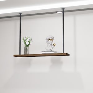 May include: A floating shelf suspended from the ceiling by black metal rods. The shelf holds a vase of white flowers, a bust of a classical figure with gold-colored glasses, and a small stack of books. The shelf is made of dark wood.