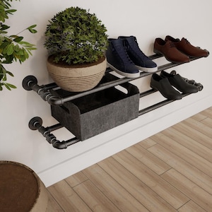 May include: A two-tiered shoe rack made from black metal pipes, mounted on a white wall. The top shelf holds two pairs of shoes and a potted plant. The bottom shelf has a gray storage bin and a pair of shoes.